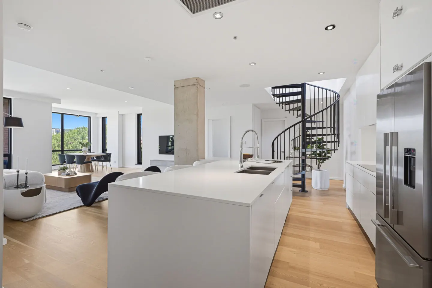 Modern open-concept kitchen with white island, stainless steel fridge, and black spiral staircase in a bright living area.