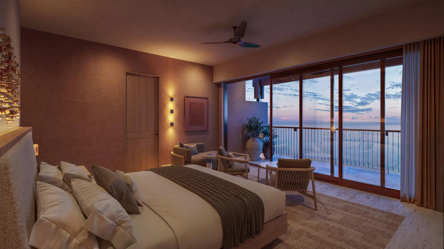 A bedroom with a king-size bed, a seating area, and a balcony with ocean views. The walls are a warm, earthy tone, and the room is lit by sconces.