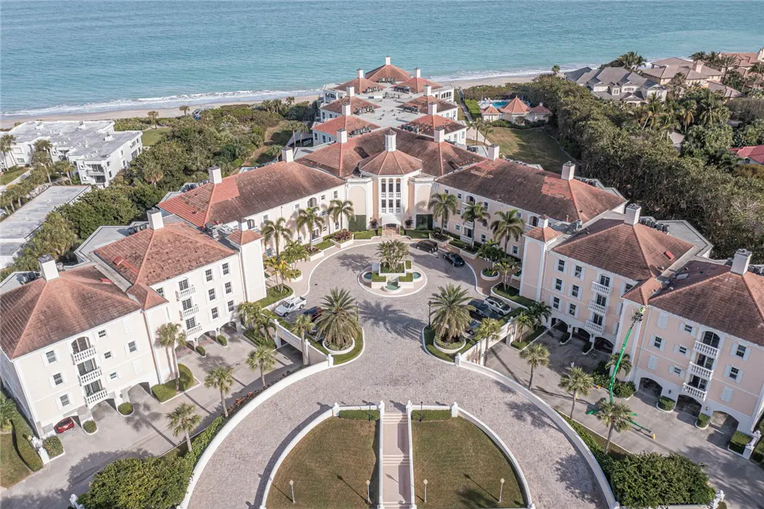 Aerial view of a beachfront resort with beige buildings, red tile roofs, palm trees, and a circular driveway. Ocean in the background.