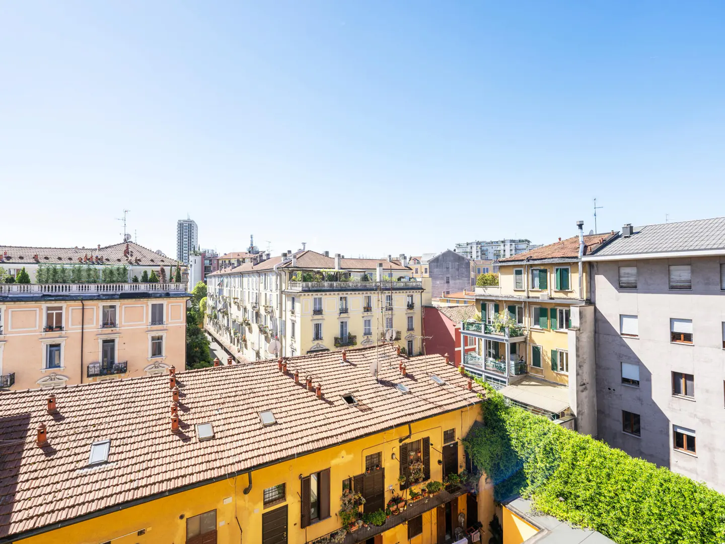 High-angle view of a sunny Italian city street with colorful apartment buildings, terracotta roofs, and clear blue sky.