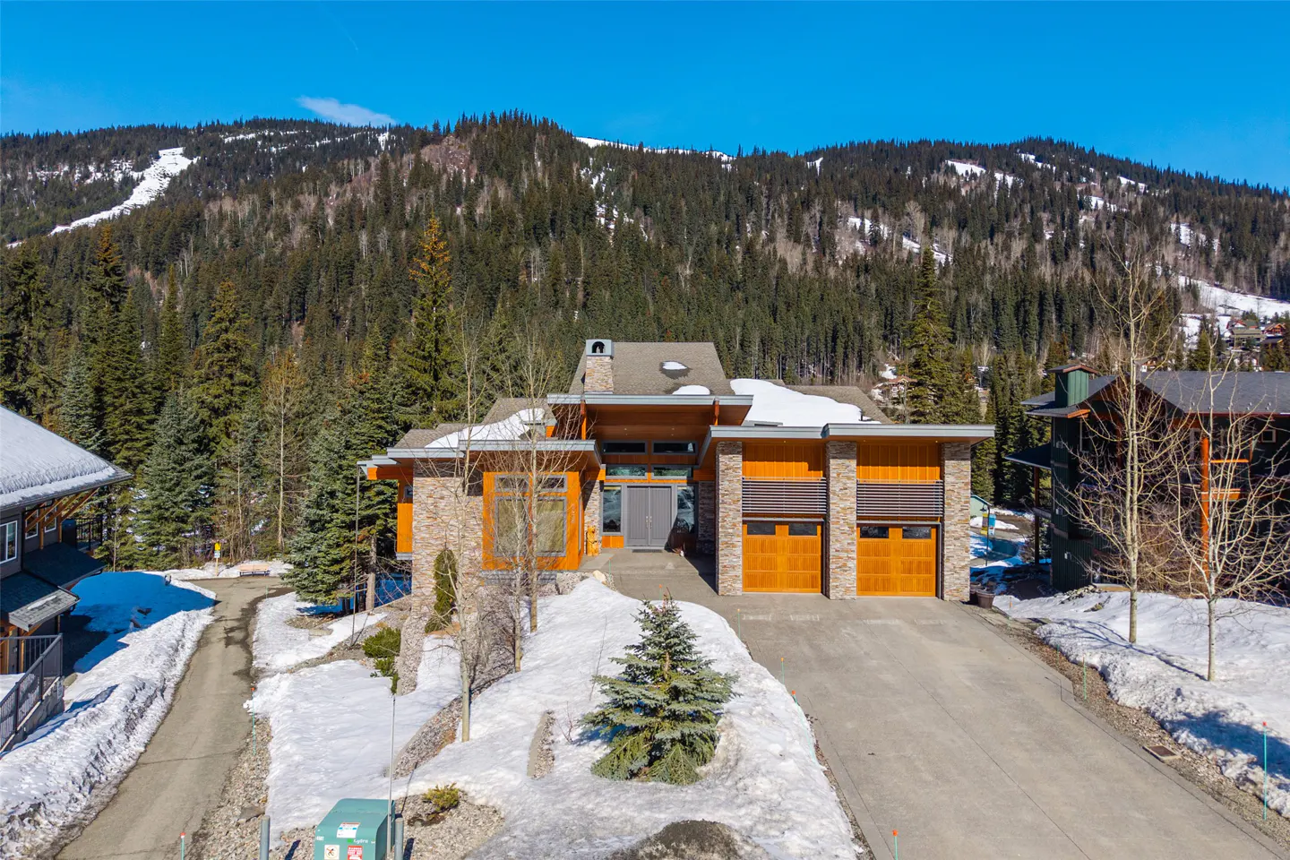 Modern mountain home with stone and wood exterior, two-car garage, and snow-covered mountain backdrop.