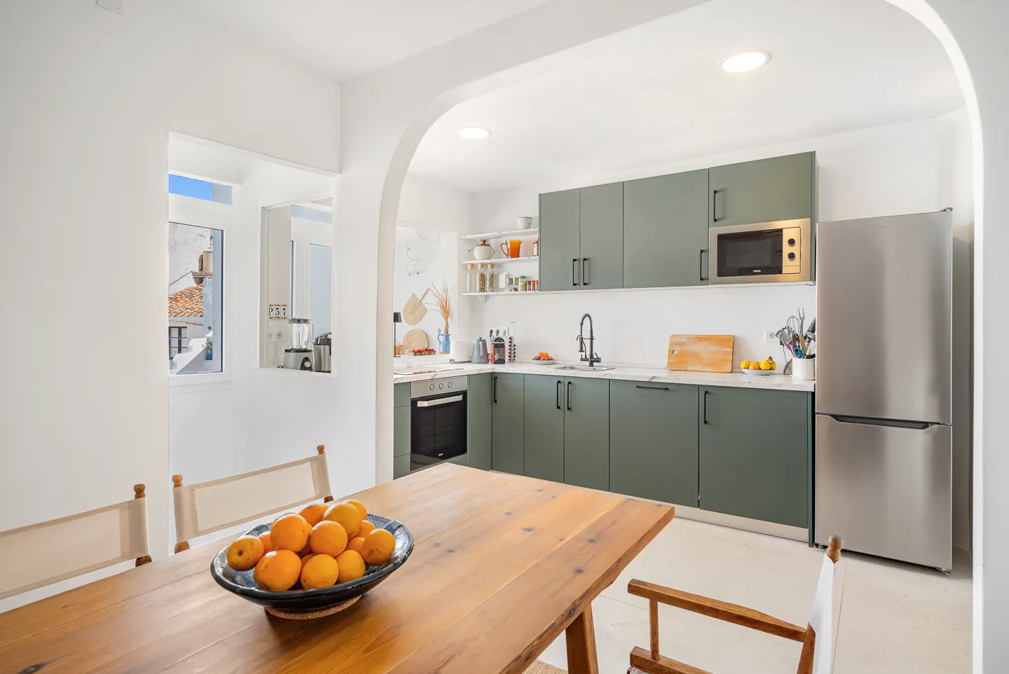 Bright kitchen with green cabinets, stainless steel fridge, and wooden table. A bowl of oranges sits on the table.