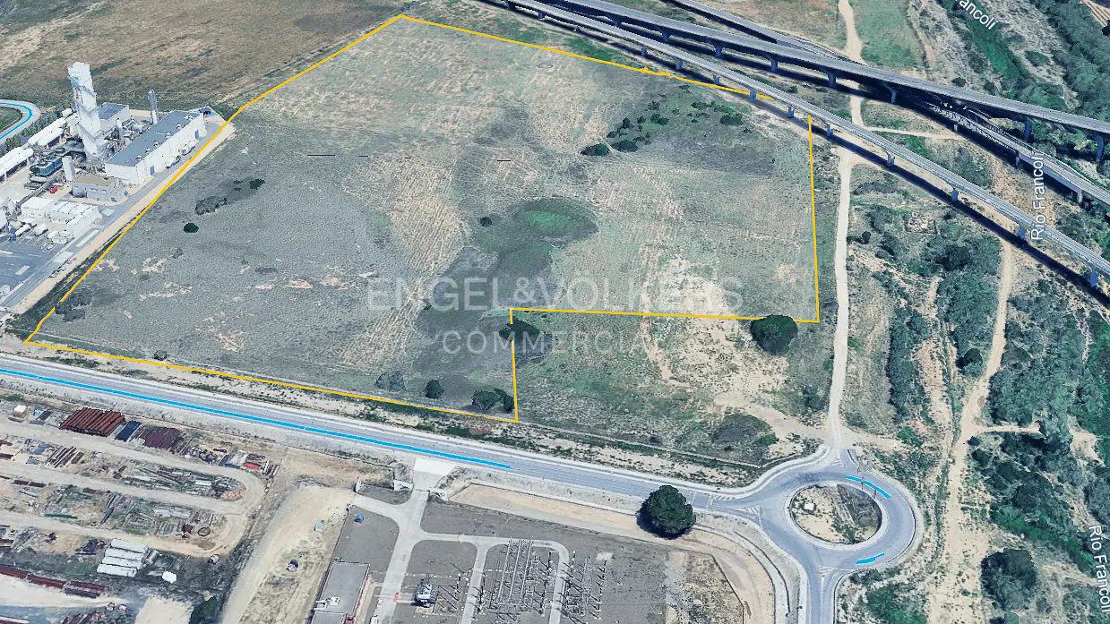 Aerial view of a vacant, yellow-outlined plot of land near an industrial facility, highway overpass, and a roundabout.