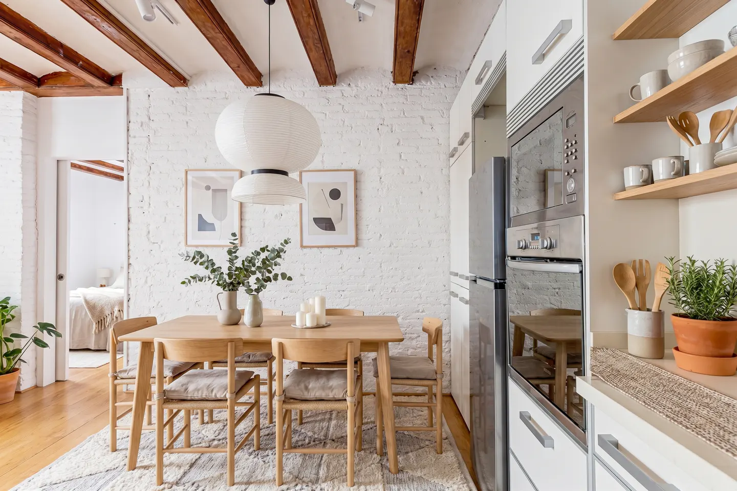 Bright kitchen with a wooden table, chairs, and a white paper lantern. White brick wall with framed art. Stainless steel appliances and open wooden shelves.