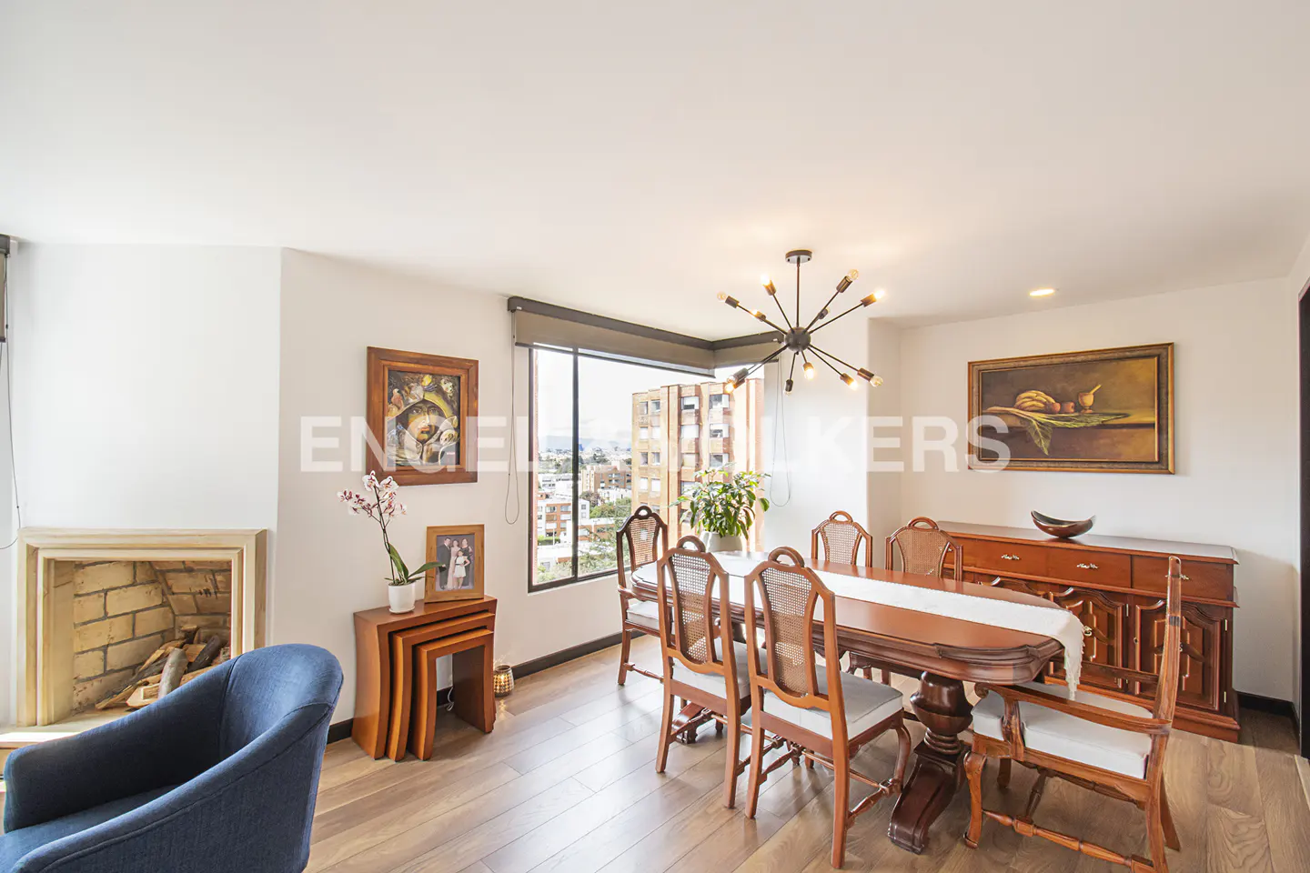 Bright dining room with wood floors, table, and chairs. A fireplace, blue chair, and city view are also visible.