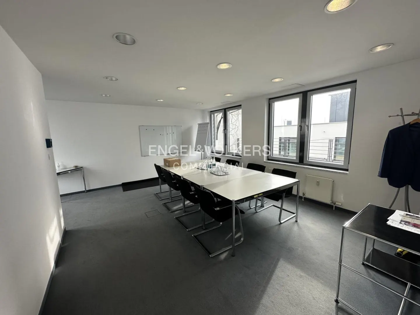 Bright conference room with a long table, black chairs, white walls, and large windows. Engel & Volkers logo visible.