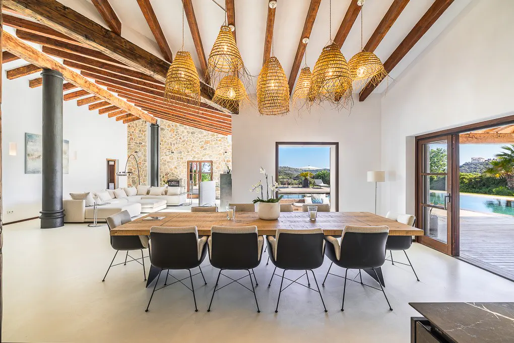 Bright, modern dining room with a long wooden table, black chairs, and woven pendant lights. Pool and landscape visible through open doors.