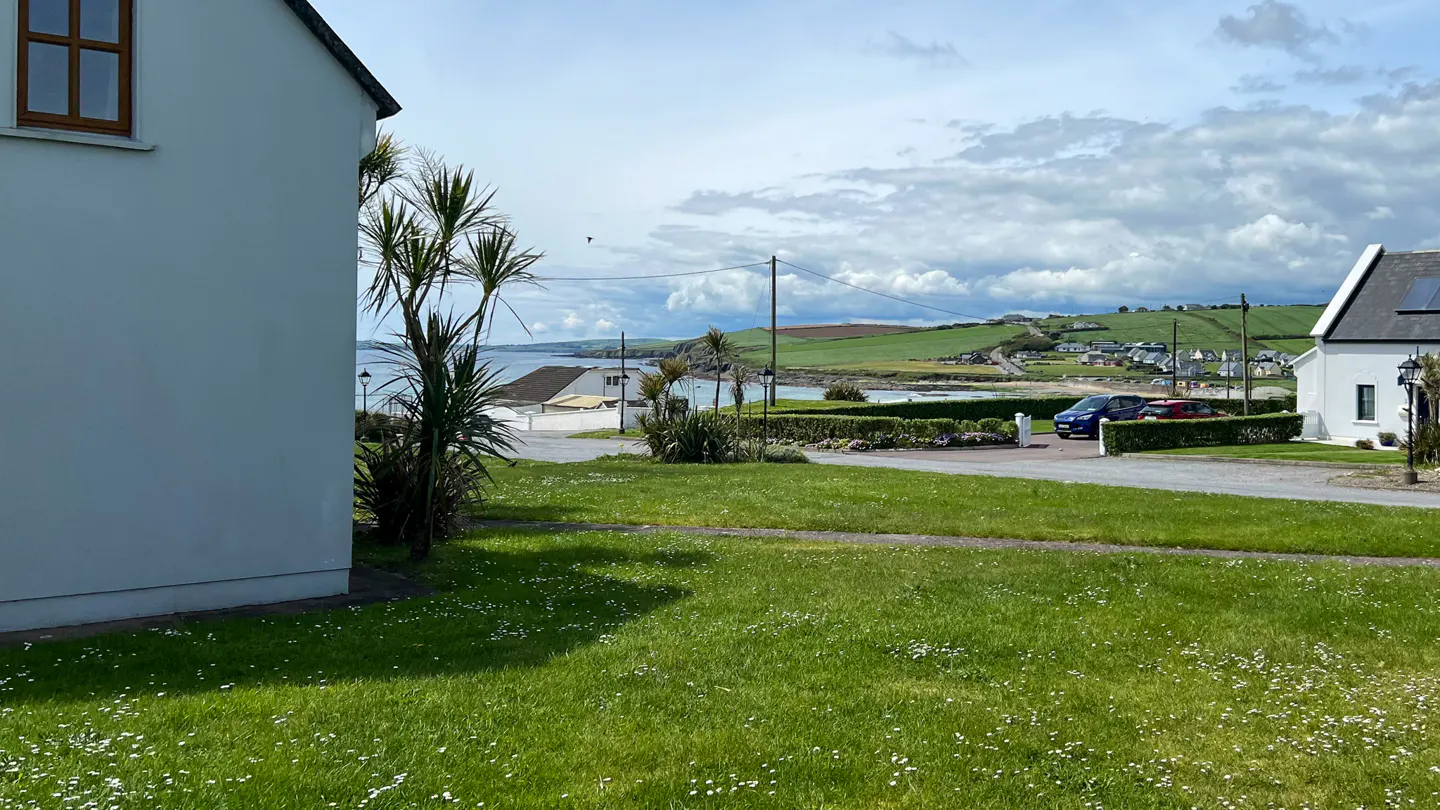 Lawn view of a white house near the coast. Green grass with white flowers in the foreground. Ocean, green hills, and cloudy sky in the background.