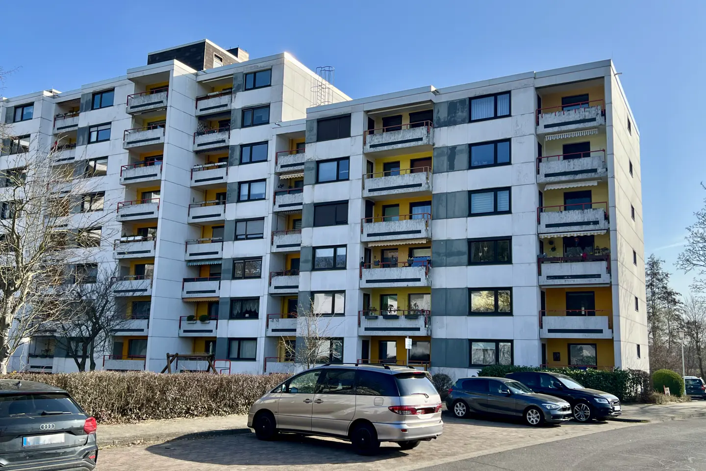 Exterior view of a tall, white apartment building with balconies and parked cars in front.