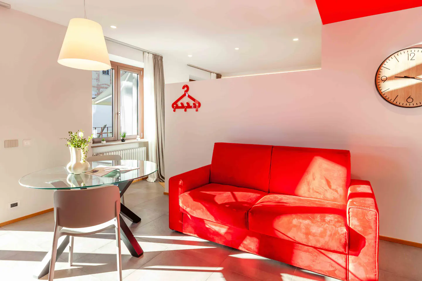 Bright living room with a red sofa, glass table with flowers, and a wooden clock on the white wall. Sunlight streams through the window.