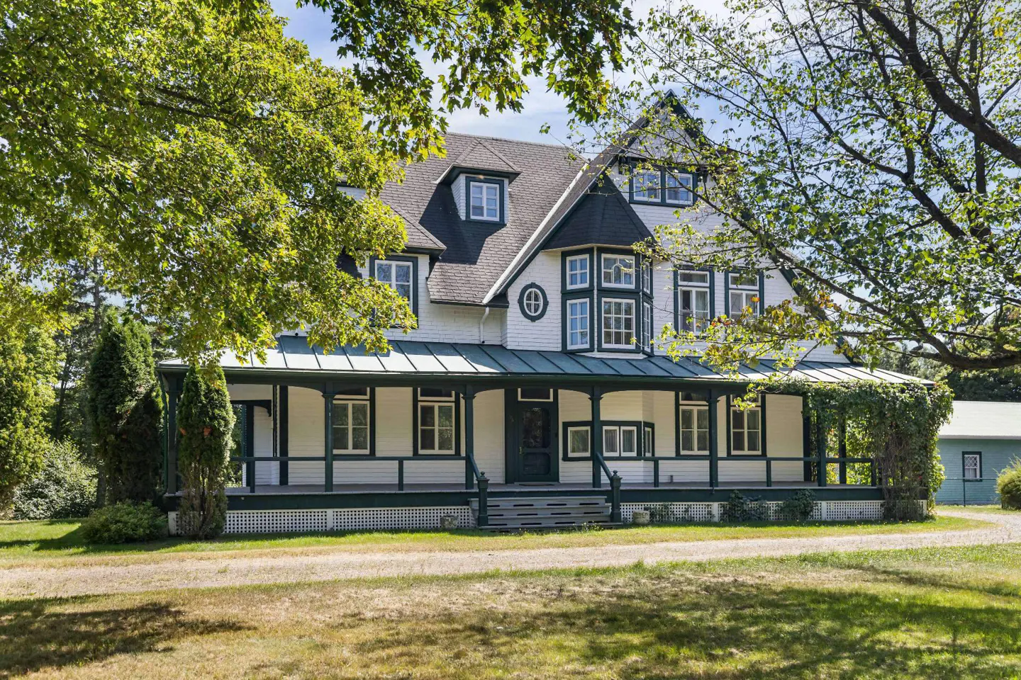 Exterior view of a large, white house with a green-trimmed porch and black roof, surrounded by trees and a green lawn.