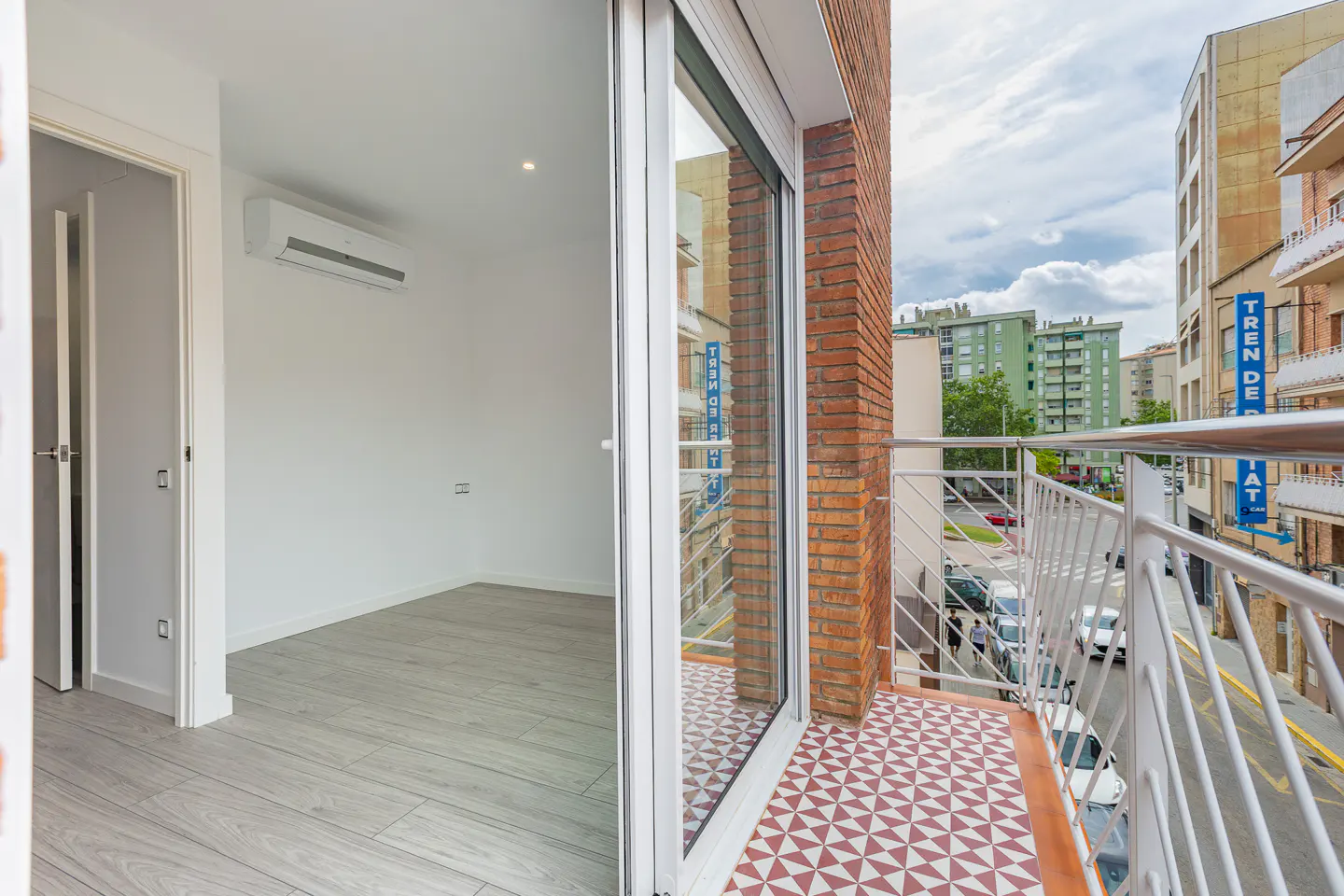 Modern room with light wood floors and AC unit, opening to a balcony with patterned tiles and city street views.