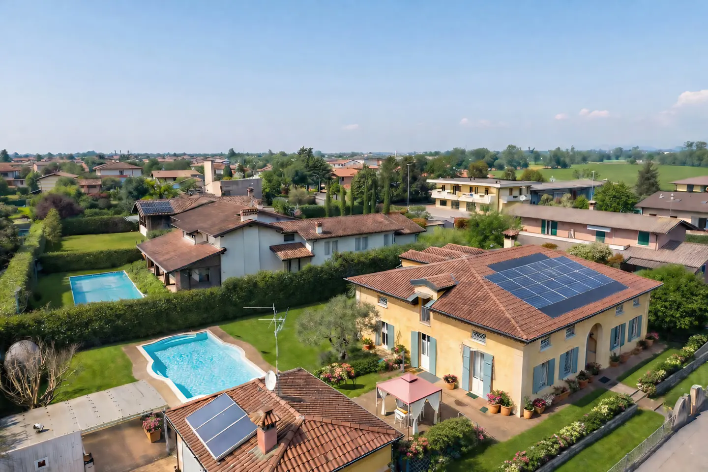 Aerial view of a yellow two-story house with blue shutters and solar panels on the roof, surrounded by green lawns and trees.