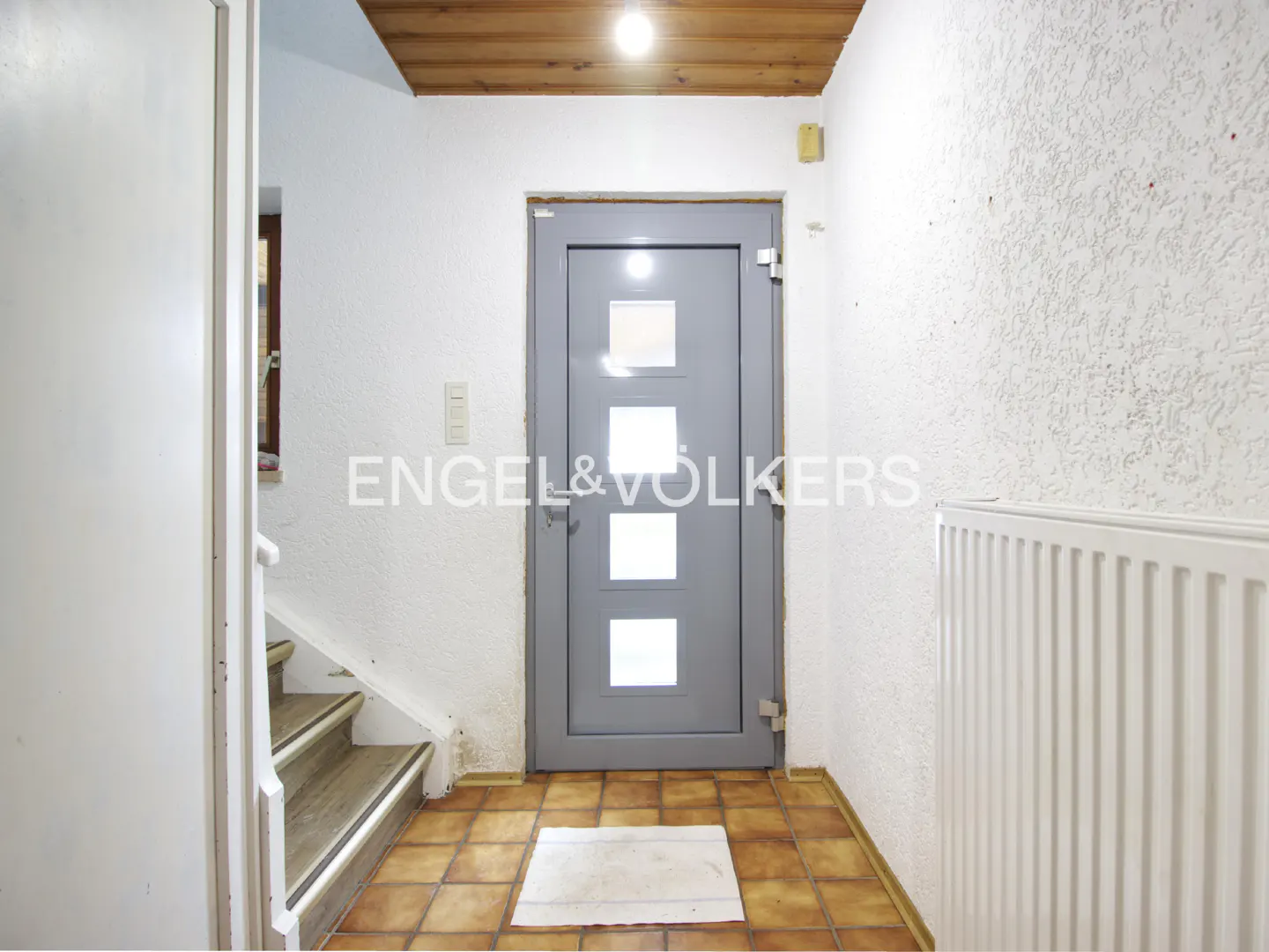 Entryway with a gray door, brown tile floor, and white walls. A staircase is visible on the left, and a radiator is on the right.