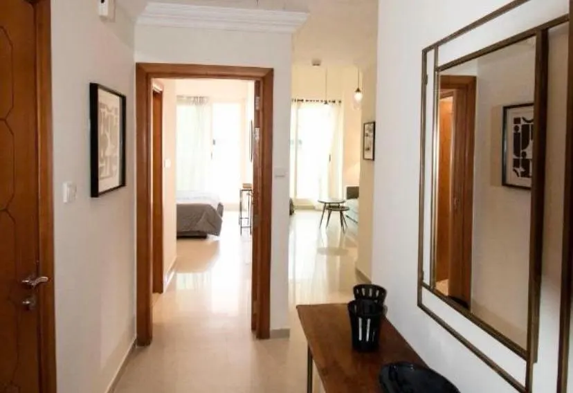 A bright hallway with a wooden console table, a large mirror, and a view into a bedroom and living area with white floors.