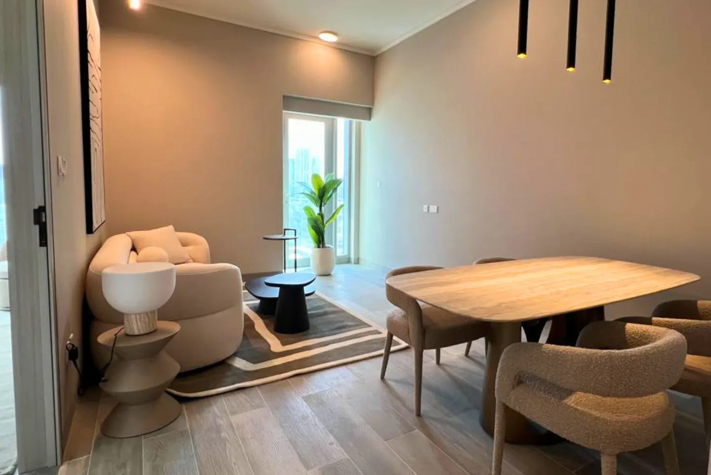 A modern living and dining area with beige walls, a light wood table with four chairs, and a cream-colored armchair. A potted plant sits near a window.