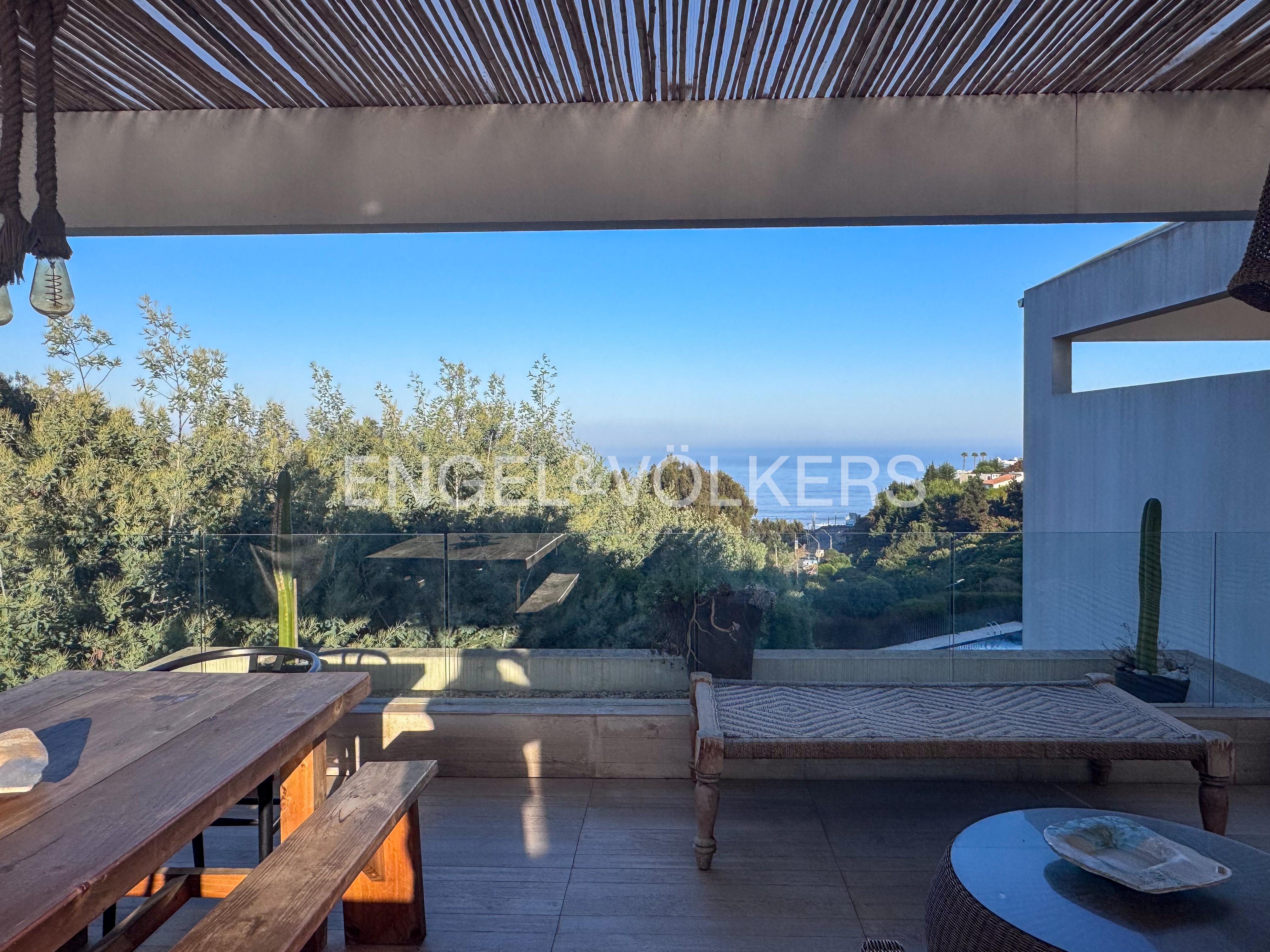 Outdoor patio with a wooden table, bench, and daybed overlooking lush greenery and the ocean under a clear blue sky.
