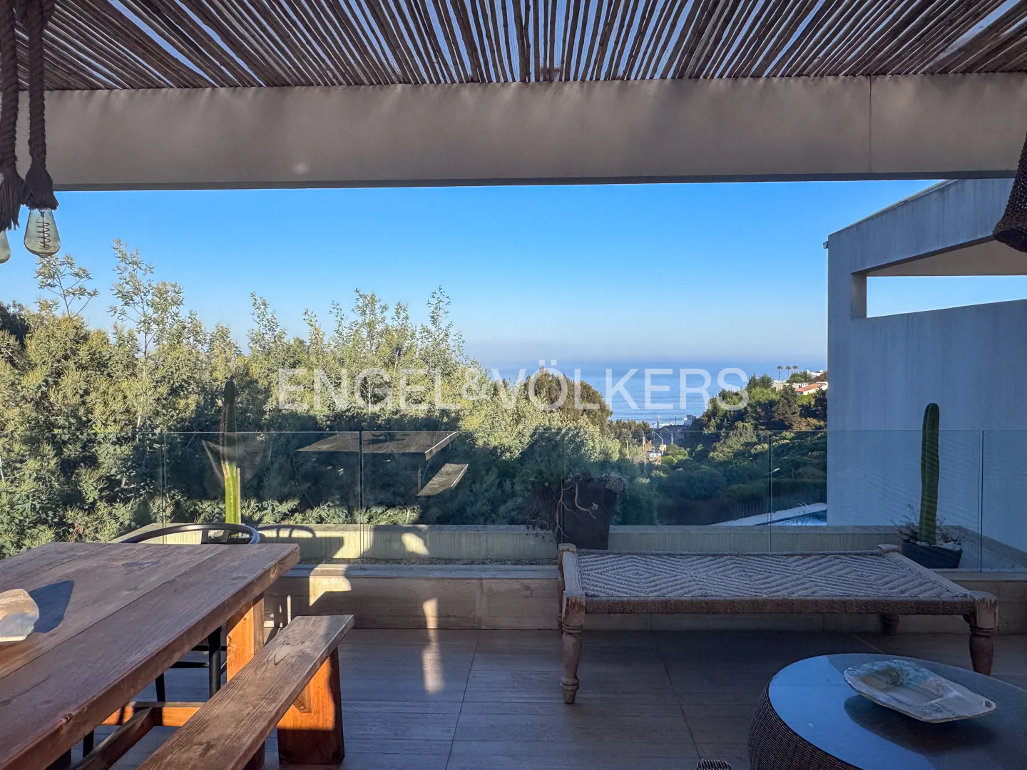 Outdoor patio with a wooden table, bench, and daybed overlooking lush greenery and the ocean under a clear blue sky.