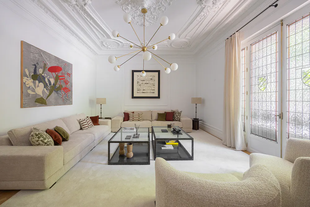 Bright living room with white walls, ornate ceiling, and a modern gold chandelier. Beige sofas and chairs surround glass coffee tables on a white rug. Artwork adorns the walls.