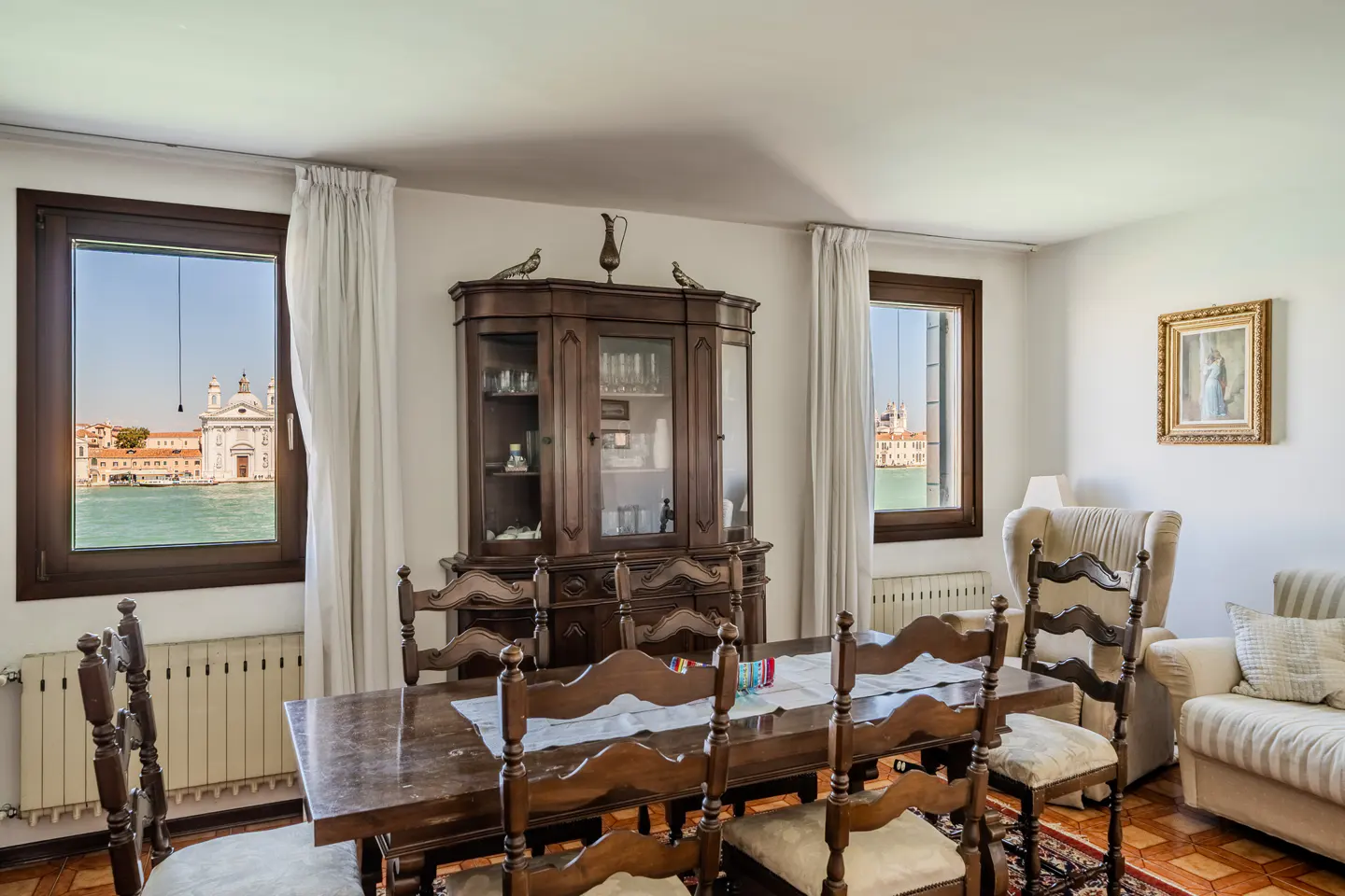 A dining room with a dark wood table and chairs, a china cabinet, and a view of Venice through two windows.
