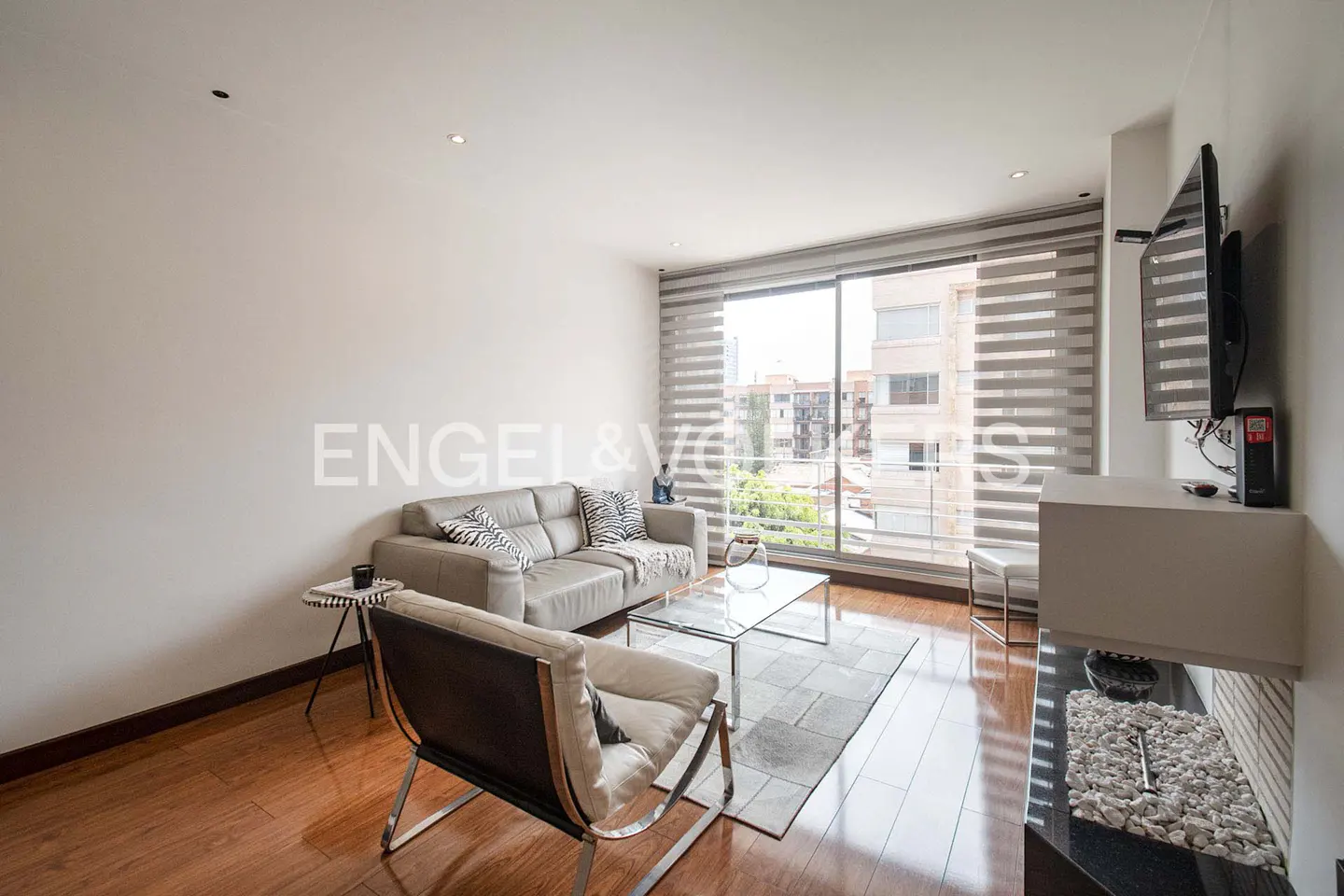 Living room with a gray sofa, chair, glass table, and wood floors. Large window with blinds and a view of a city building. TV on the wall.