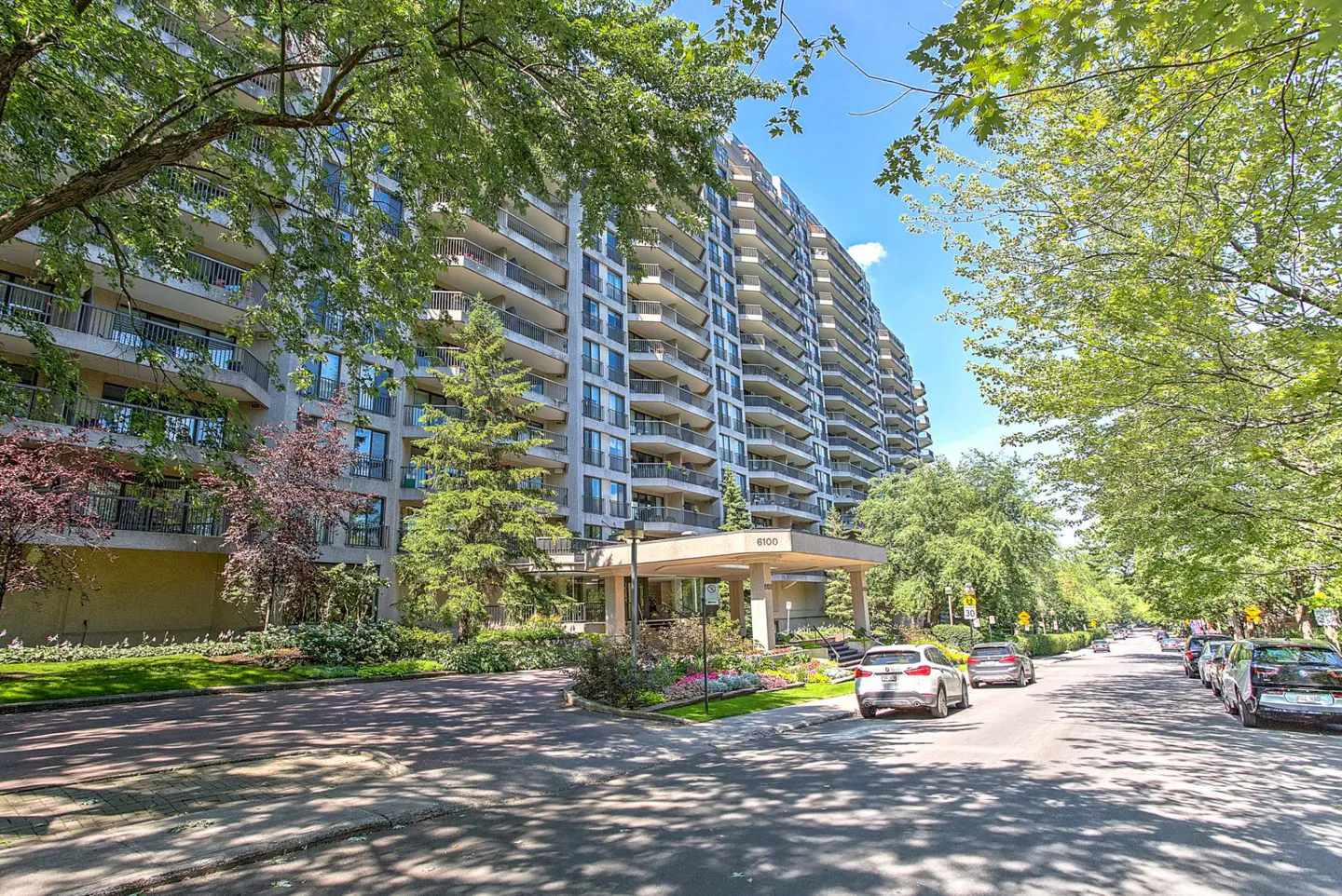 Exterior view of a tall, light gray apartment building with balconies, trees, and cars parked on the street.