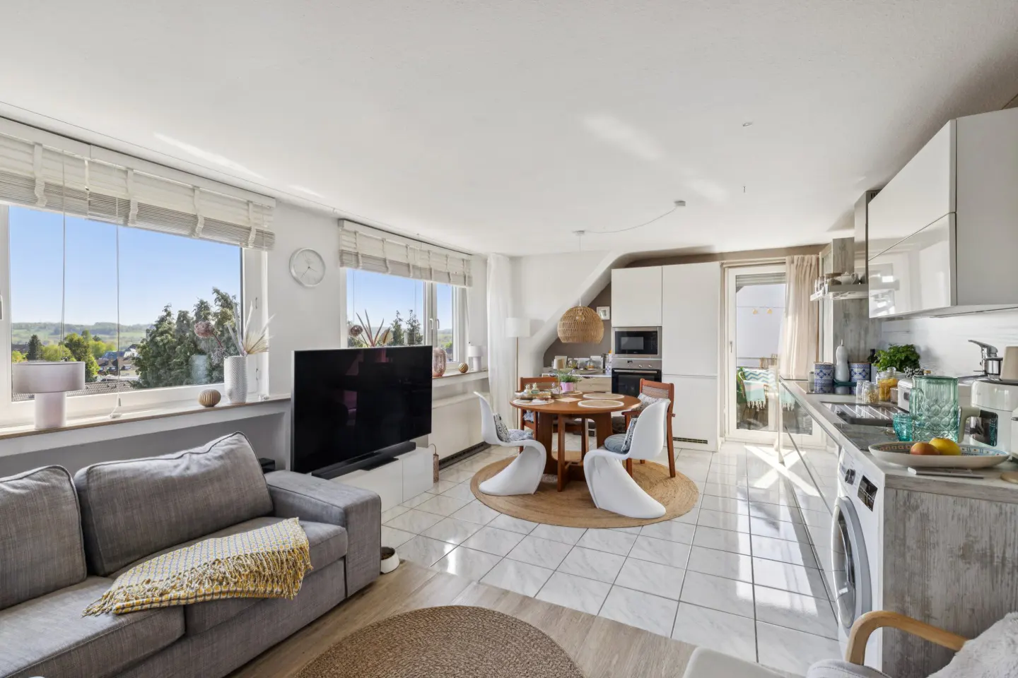 Bright, open-concept apartment with gray sofa, TV, round dining table with four chairs, and a modern white kitchen. Large windows offer a view of trees.