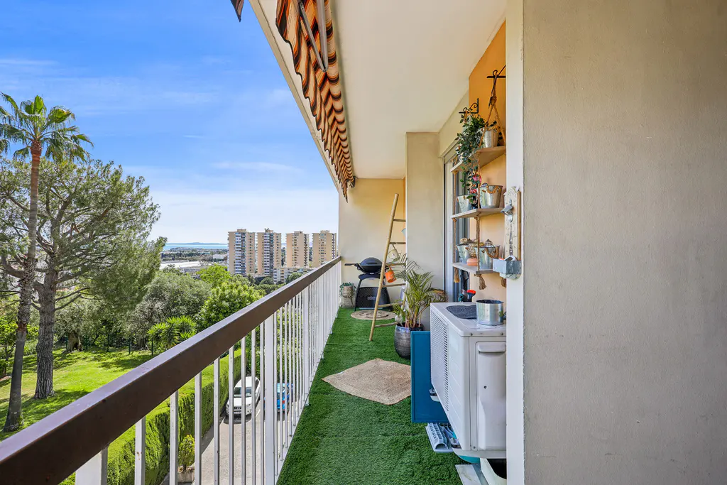 Balcony view with green turf, white railings, and a brown awning. A grill, ladder, and shelves with plants decorate the space. City buildings are visible in the distance.