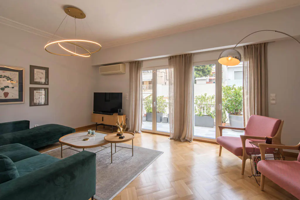 Living room with a green velvet sofa, pink chairs, and a balcony with plants. A modern chandelier hangs above a coffee table.