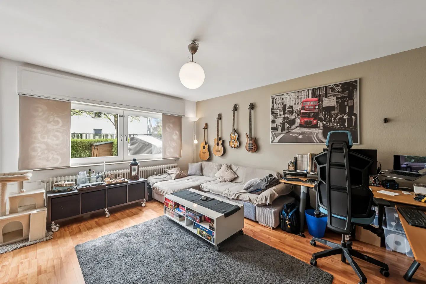 Living room with a gray sectional sofa, guitars on the wall, and a London-themed picture. A desk and chair are on the right.