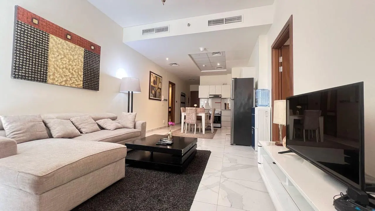 A bright apartment living room with a beige sofa, black coffee table, and a TV. The kitchen and dining area are visible in the background.