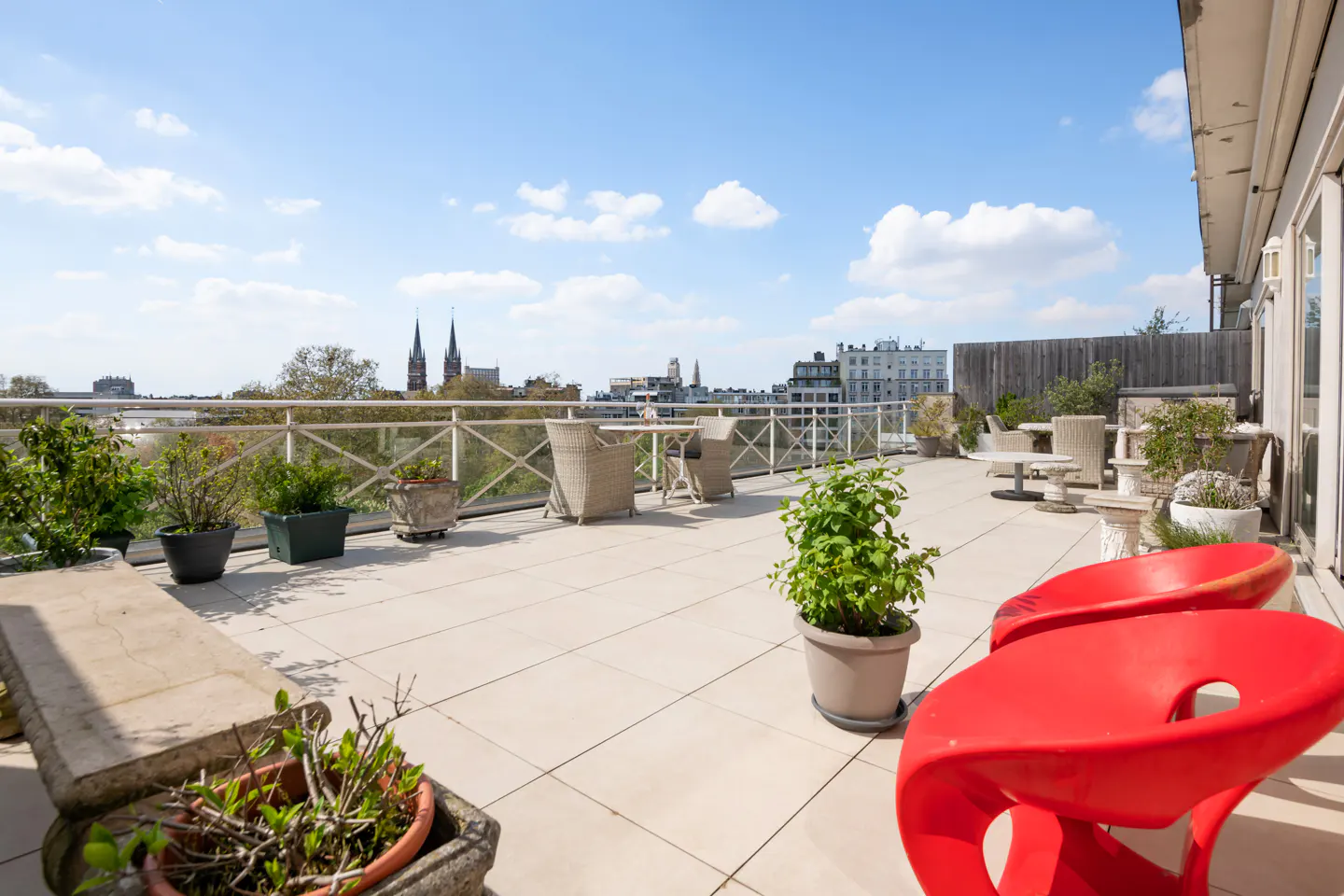 Rooftop patio with red chairs, potted plants, and city views under a blue sky with white clouds.