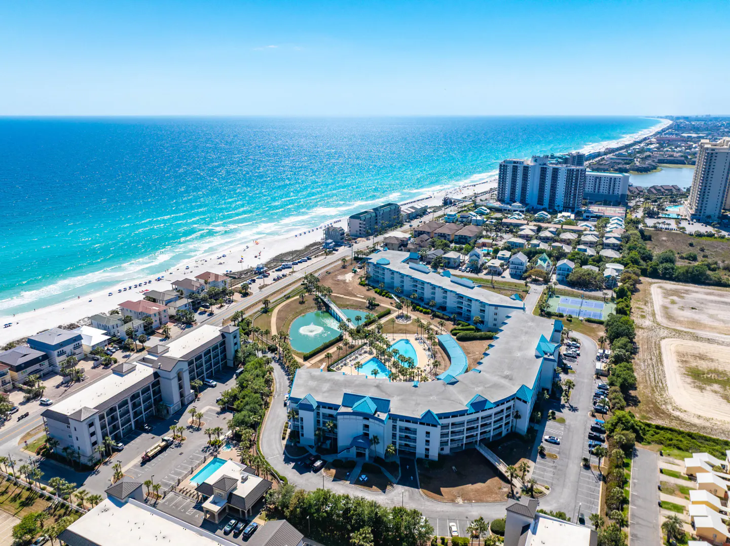 Aerial view of a beach resort with turquoise water, white sand, and blue-roofed buildings under a clear blue sky.