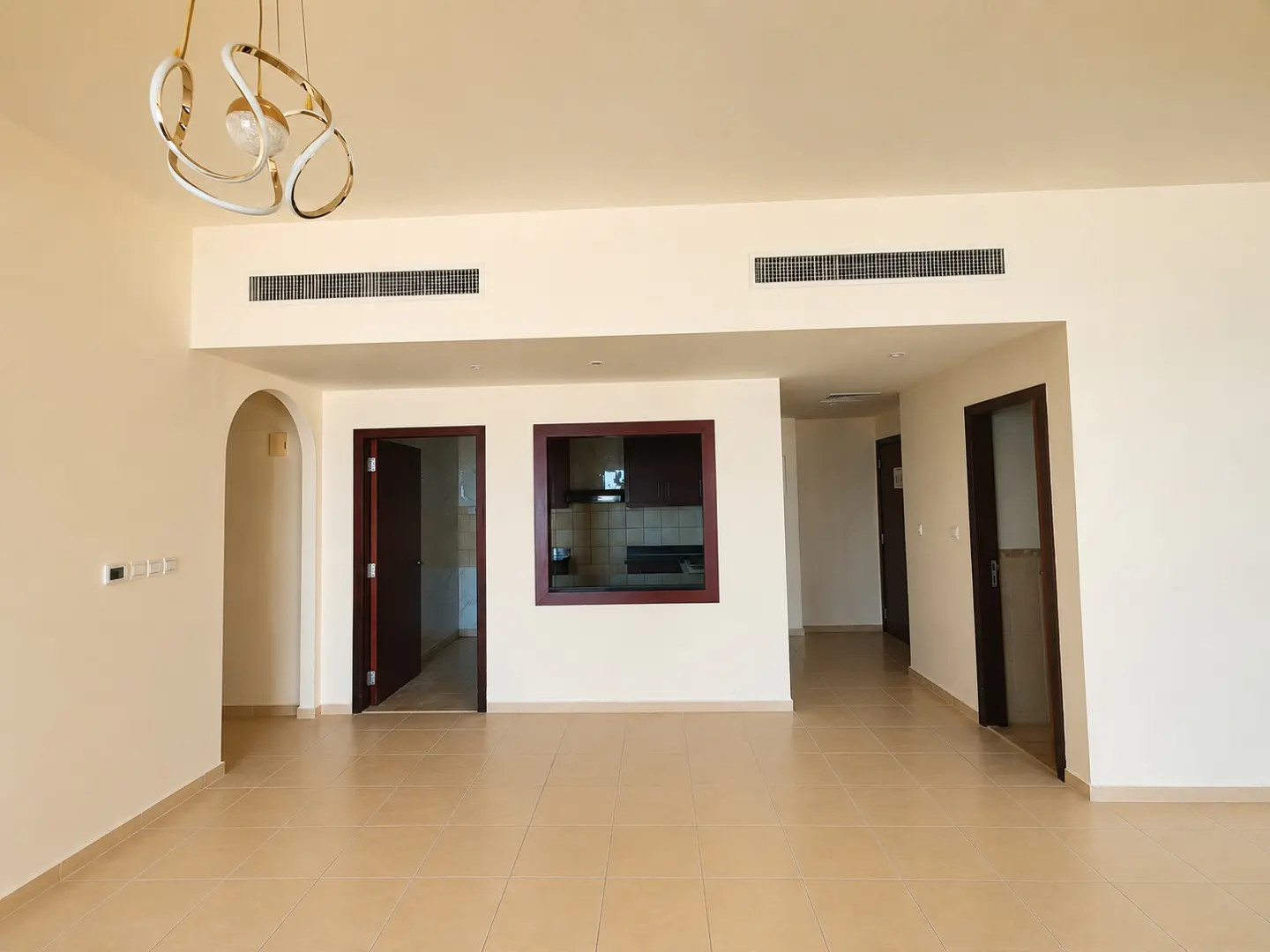 Empty living room with beige walls and tile floor. A modern chandelier hangs from the ceiling. Doors and a kitchen window are visible.