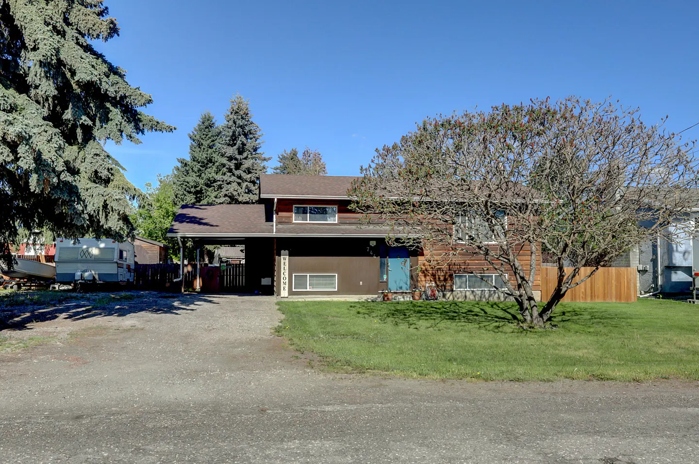 Two-story brown house with a blue door, a carport, and a "Welcome" sign, set against a clear blue sky. A large tree stands in the front yard.