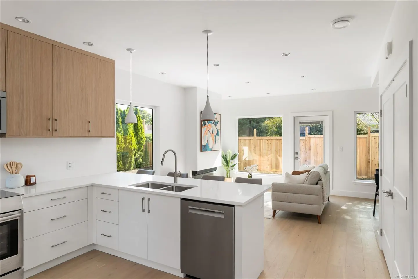 Bright, open-concept kitchen and living room with white cabinets, stainless steel appliances, and light wood floors. Windows overlook a fenced backyard.