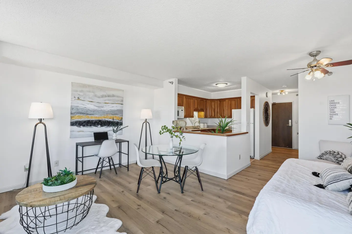 Bright, open-concept living space with wood floors, white walls, and a view into the kitchen. A desk, table, and sofa furnish the room.