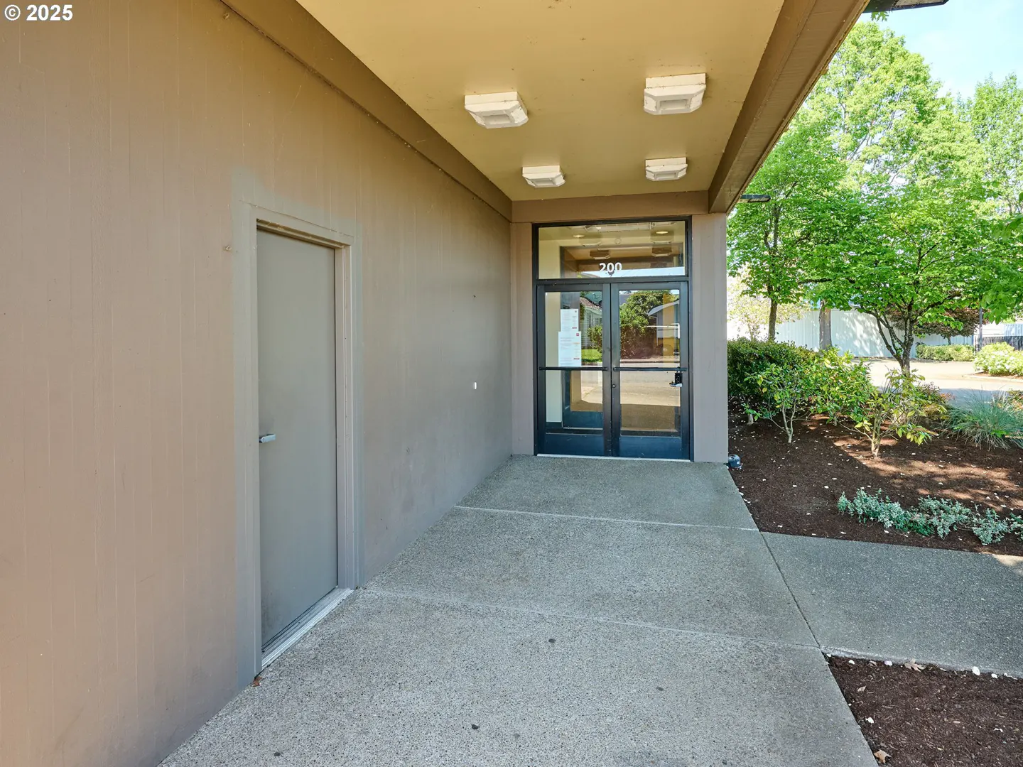 Exterior view of a tan building with a glass door entrance marked "200" and a gray side door, under a covered walkway with square lights.