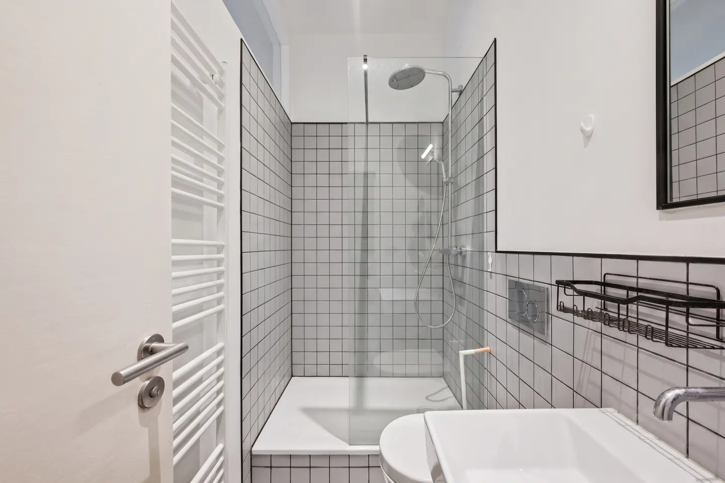 Modern bathroom with white square tiles, black trim, glass shower, white sink, and towel rack.