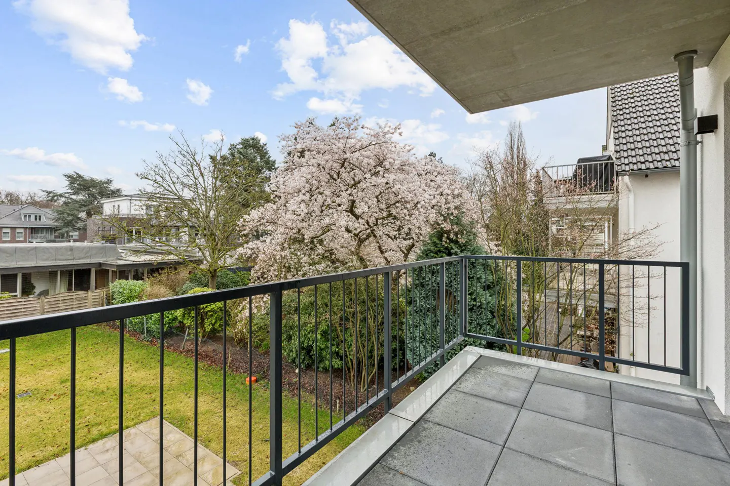 Balcony view with black railings overlooking a green lawn and flowering tree under a blue, cloudy sky.