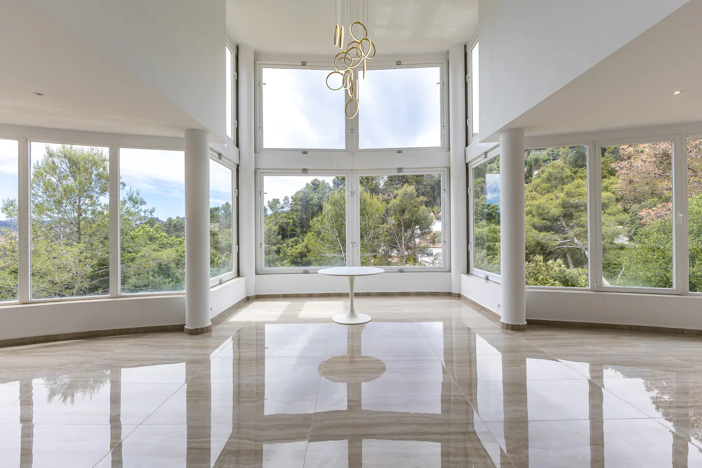 Bright, empty room with marble floors, white walls, and large windows showing trees. A white table sits in the center, with a modern chandelier above.