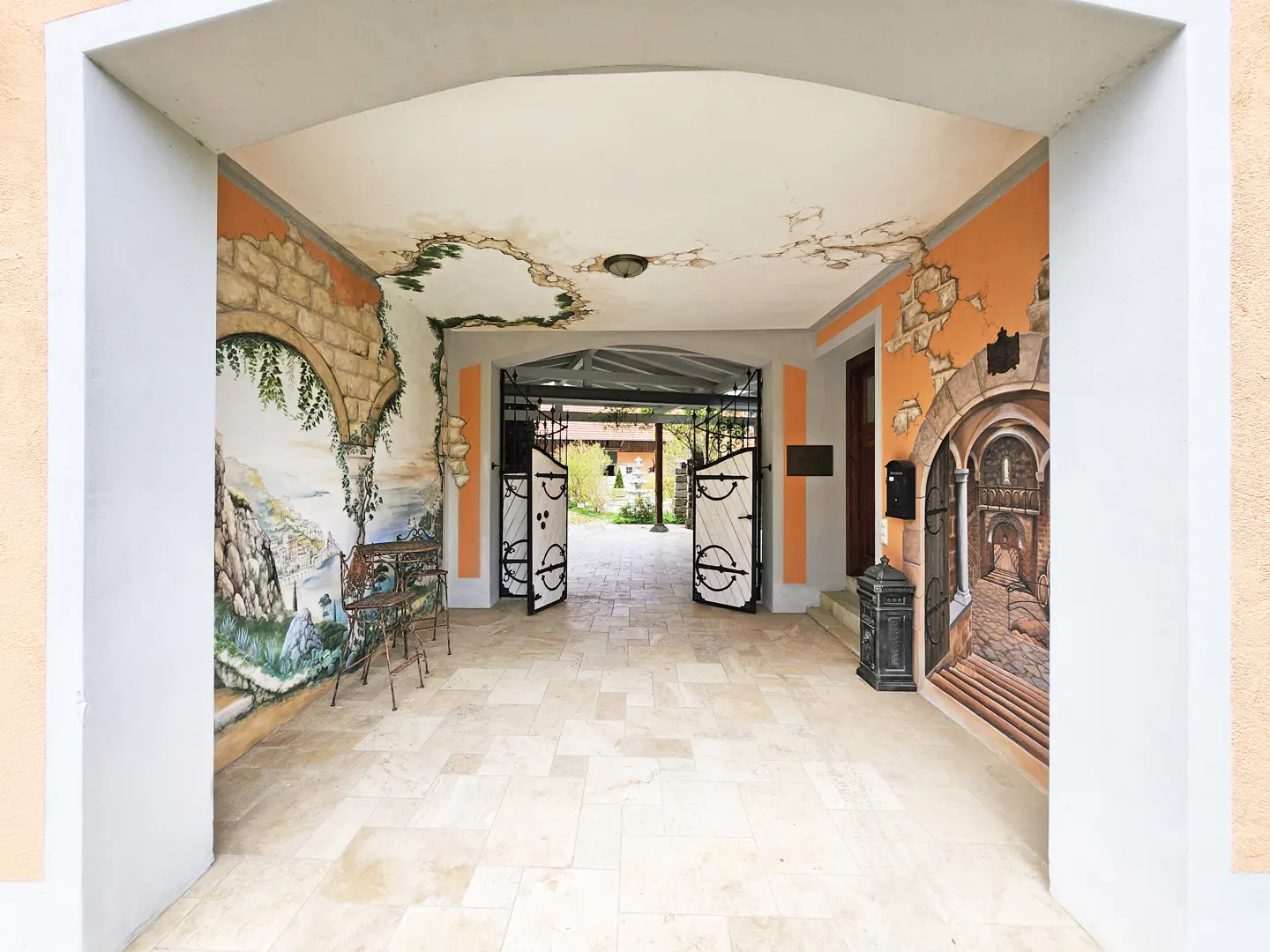 Covered entryway with painted murals on orange walls, leading to an open black metal gate and garden.