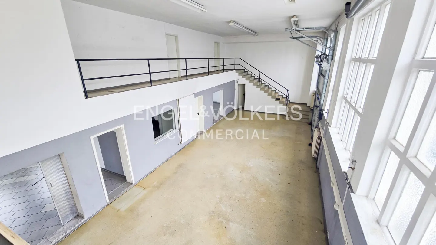 Bright, empty commercial space with a mezzanine, stairs, and large windows. Walls are white and gray. "Engel & Völkers Commercial" logo visible.