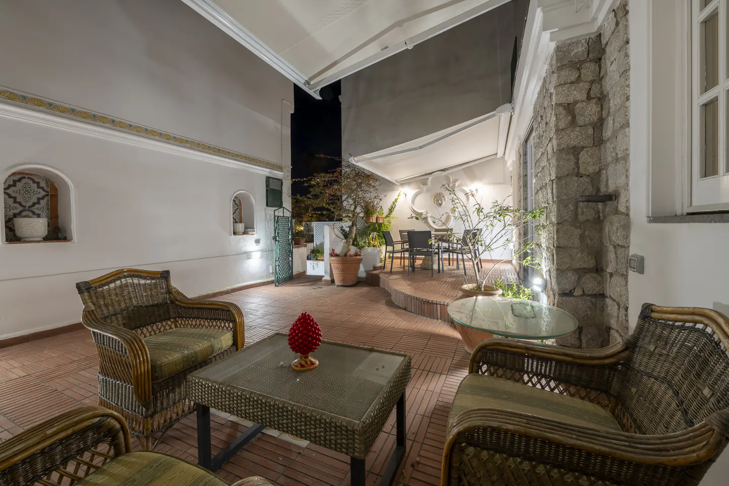Outdoor patio with wicker furniture, a glass table, and a red pinecone decoration. The patio has a stone wall and a white wall with arched niches.