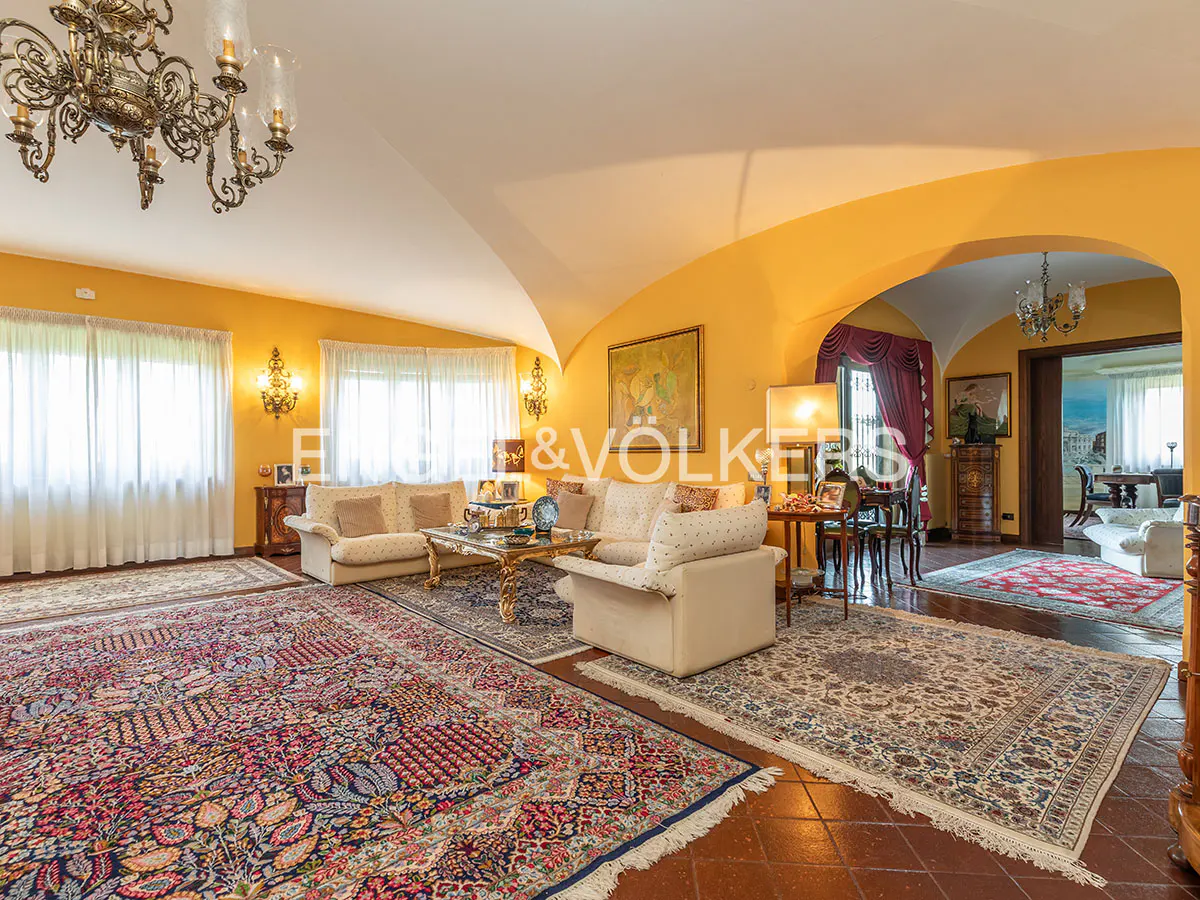 A bright living room with yellow walls, ornate rugs, and cream sofas. A chandelier hangs from the ceiling. The Engel & Völkers logo is visible.