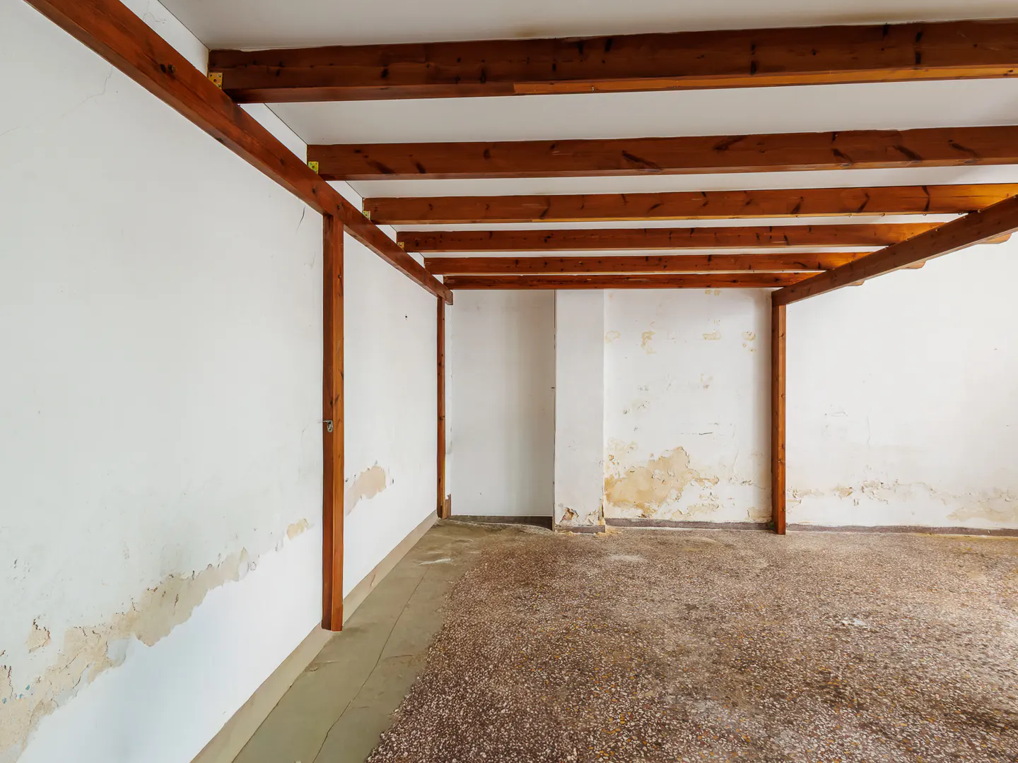 Empty room with exposed wooden ceiling beams, white walls showing signs of wear, and a textured concrete floor.
