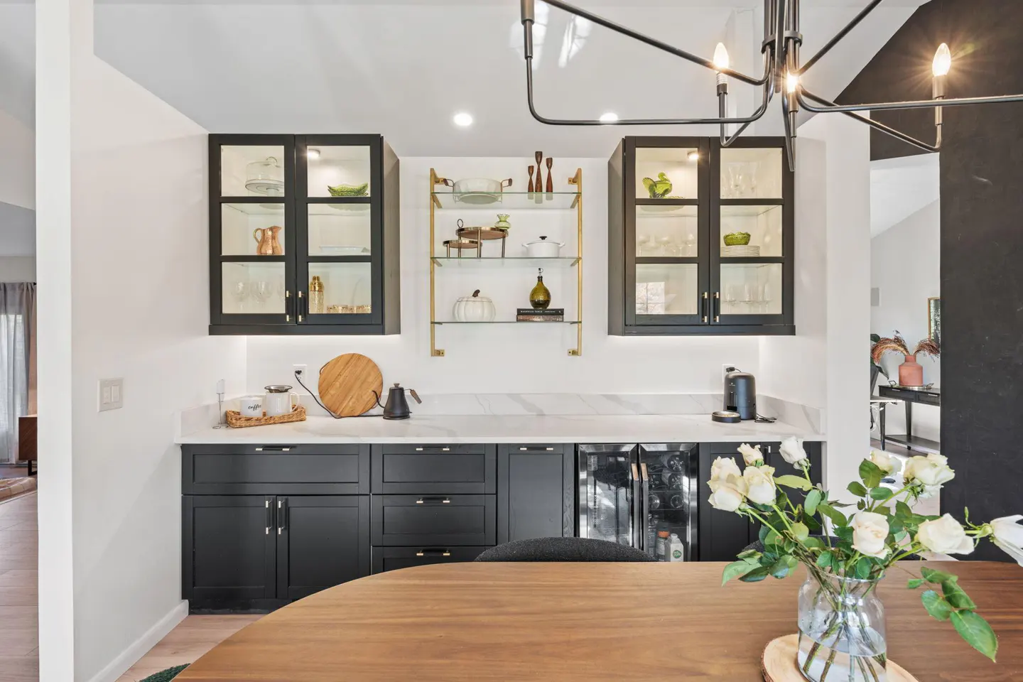 A modern kitchen with black cabinets, white marble countertops, and glass shelving. A wooden table with white roses in the foreground.