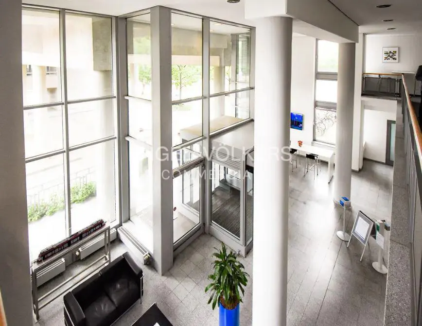 Interior view of a modern office lobby with large windows, gray tile floors, and white columns. A black sofa and green plant add color.