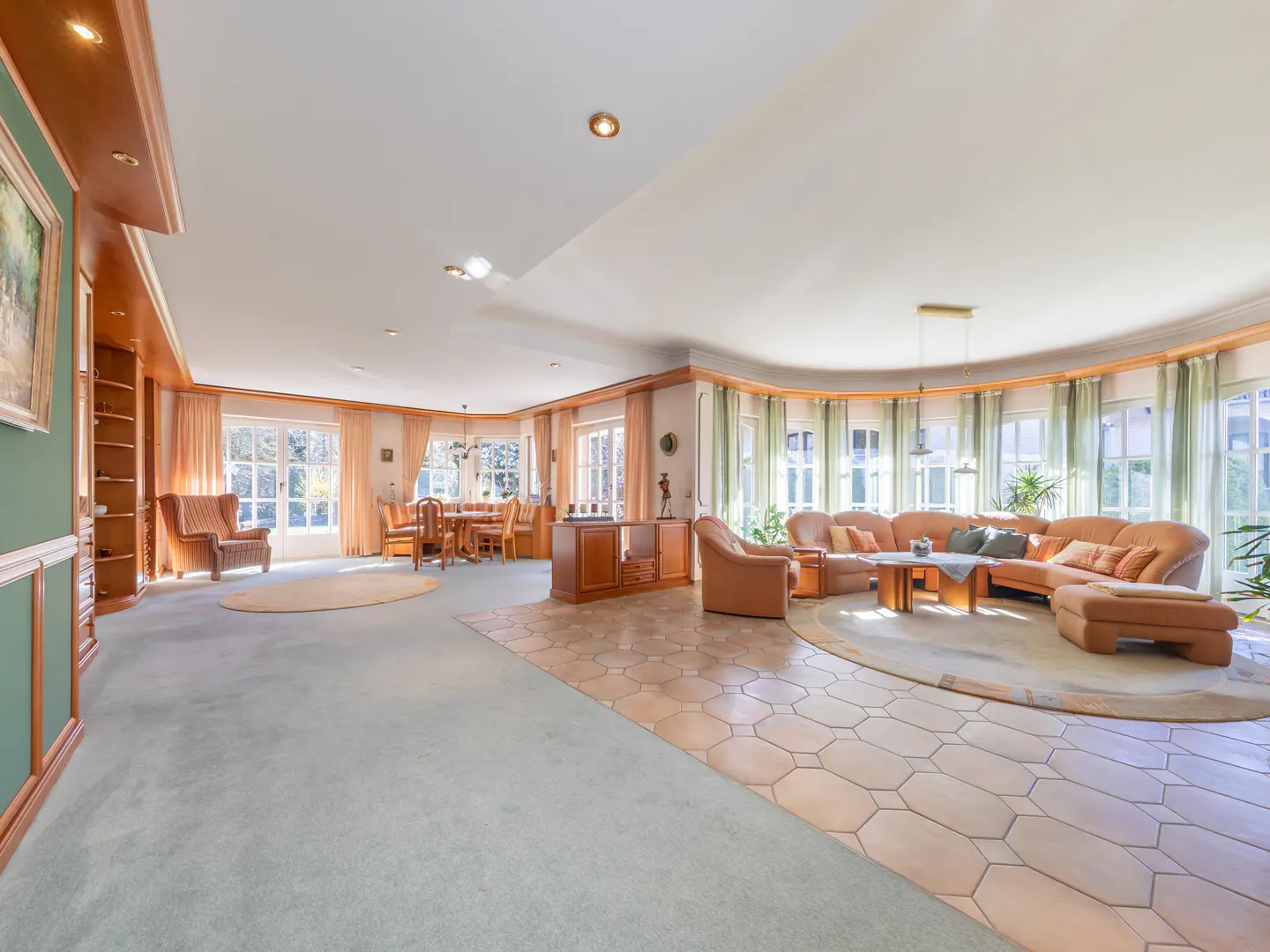 A spacious living room with a dining area, featuring a large brown sofa, a wooden dining table, and light blue carpet.
