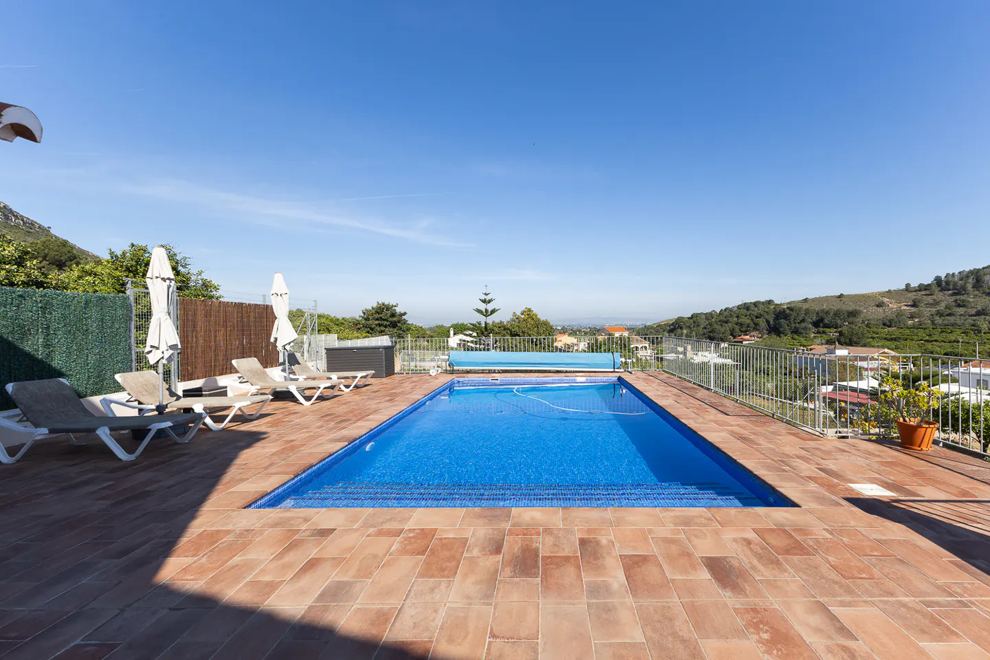 Outdoor pool with blue water and a blue cover, surrounded by terracotta tiles, lounge chairs, and a view of green hills under a clear blue sky.